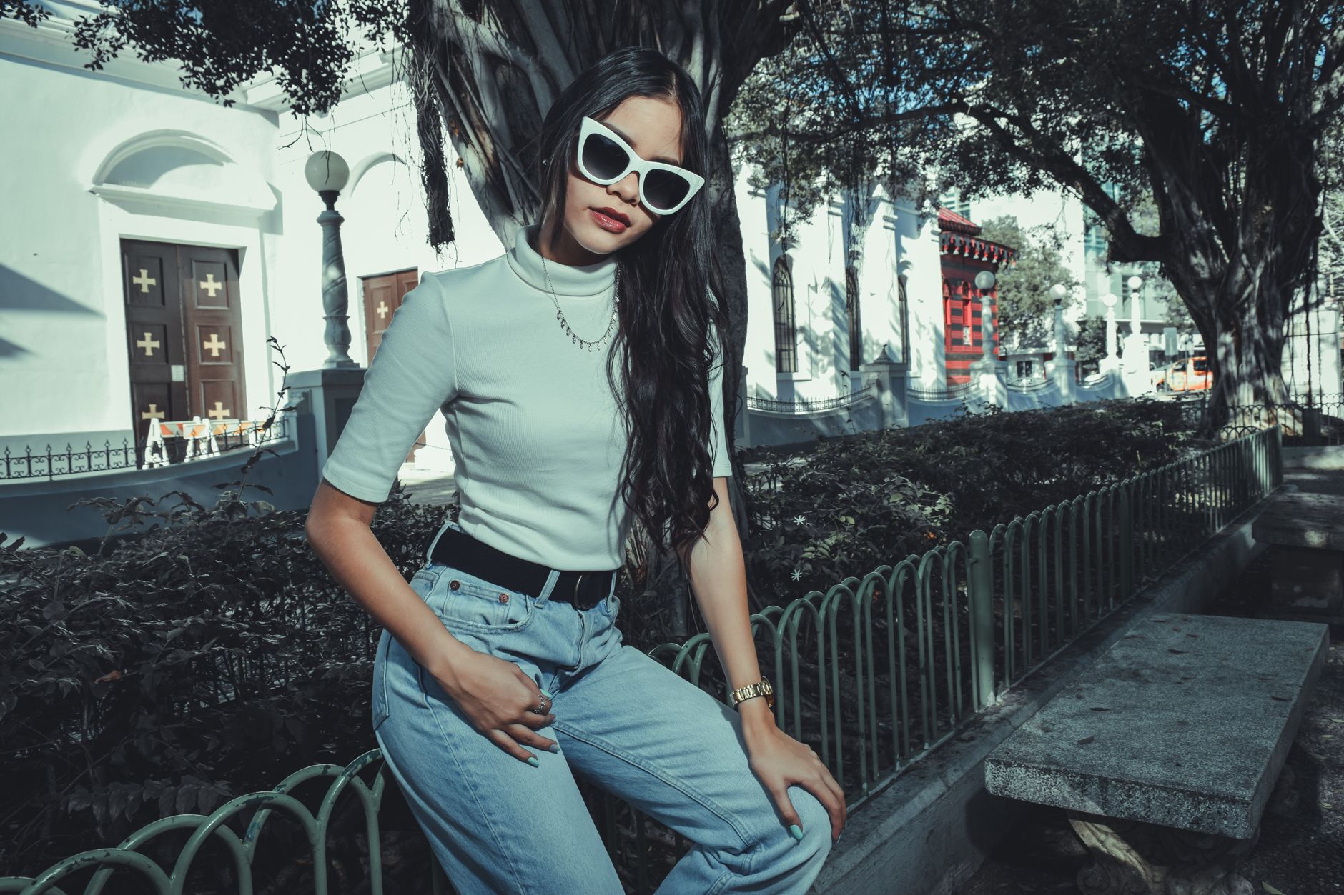 Chic woman wearing sunglasses poses by a metal fence in an urban street setting.